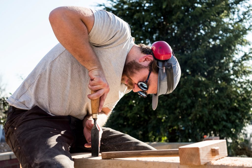 Muž v baseballové čepici, slunečních brýlích a s chrániči uší na staveništi pracuje na dřevěném trámu.