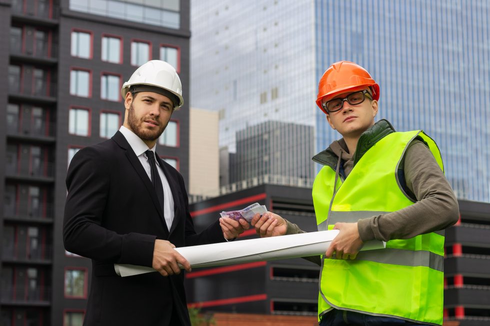 Dva muži ve stavebních helmách společně drží v rukách plány staveb a peníze, které si předávají.