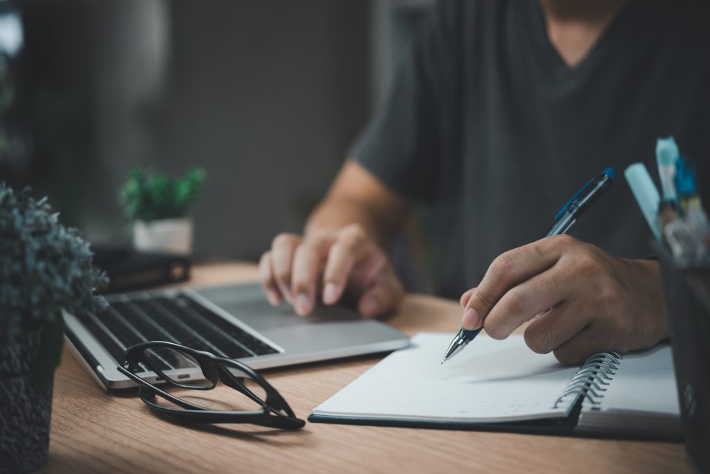 Muž píše perem na notebook v kanceláři, zaznamenává cíle, plány a úkoly. Scéna symbolizuje učení, vzdělávání a pracovní proces.